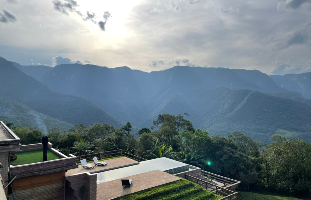 Picture of a mountain range. The mountains are covered with trees, the sky is cloudy but with enough gaps for the sun to come through. In the foreground, a pool and two chairs overlook the cliffs.